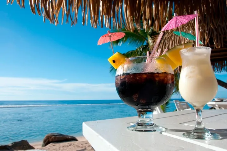 Two tropical drinks on a table by the sea, under a thatched umbrella