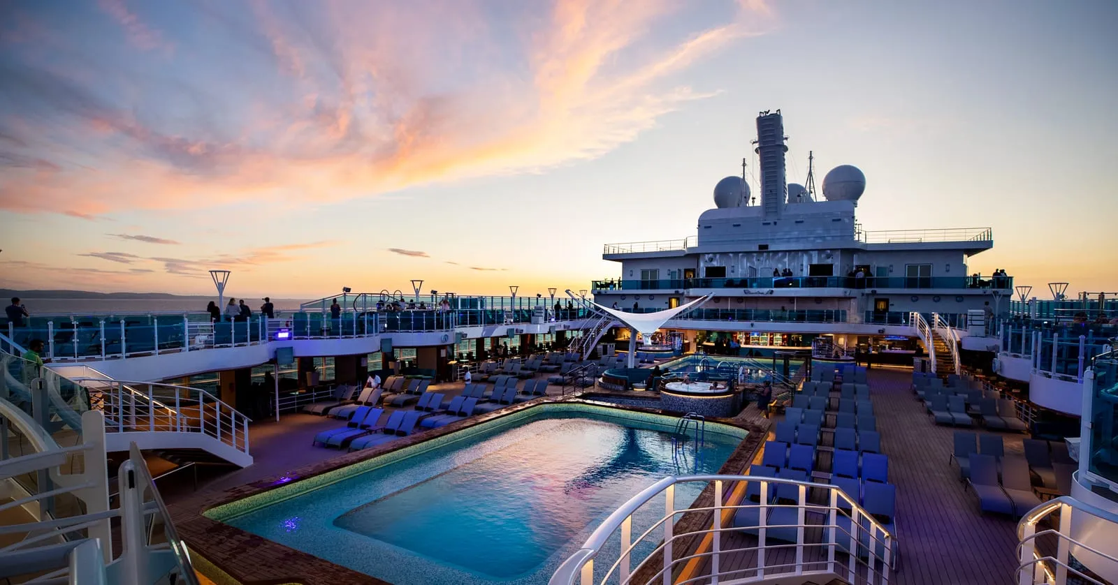 The open pool deck of a cruise ship at sunset, empty loungers and curved white railings in the foreground and flat open sea beyond.