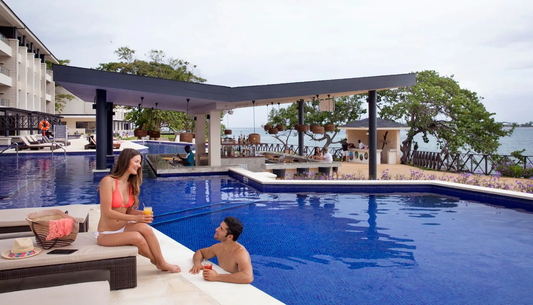 A couple relaxing at a resort pool beside a swim-up bar, with the sea beyond
