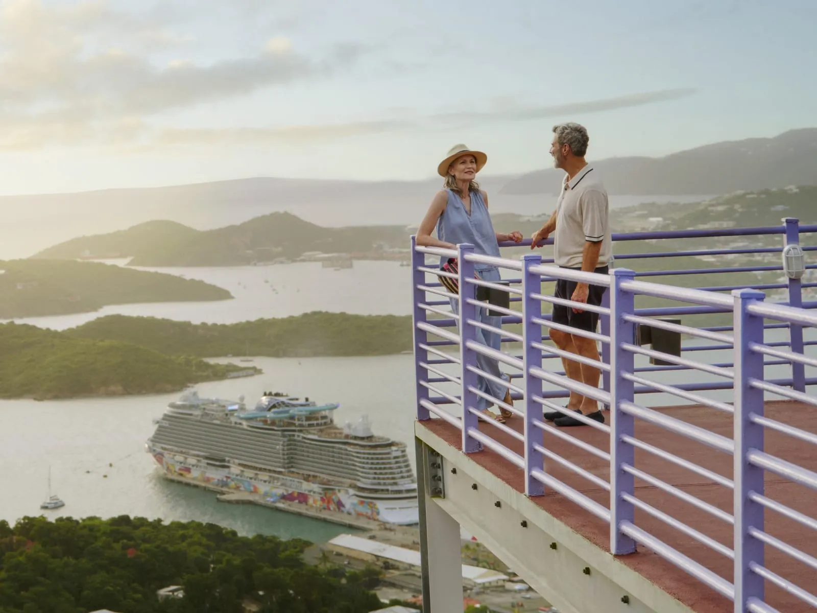 A couple at a hilltop lookout above a Caribbean harbor with a cruise ship docked below