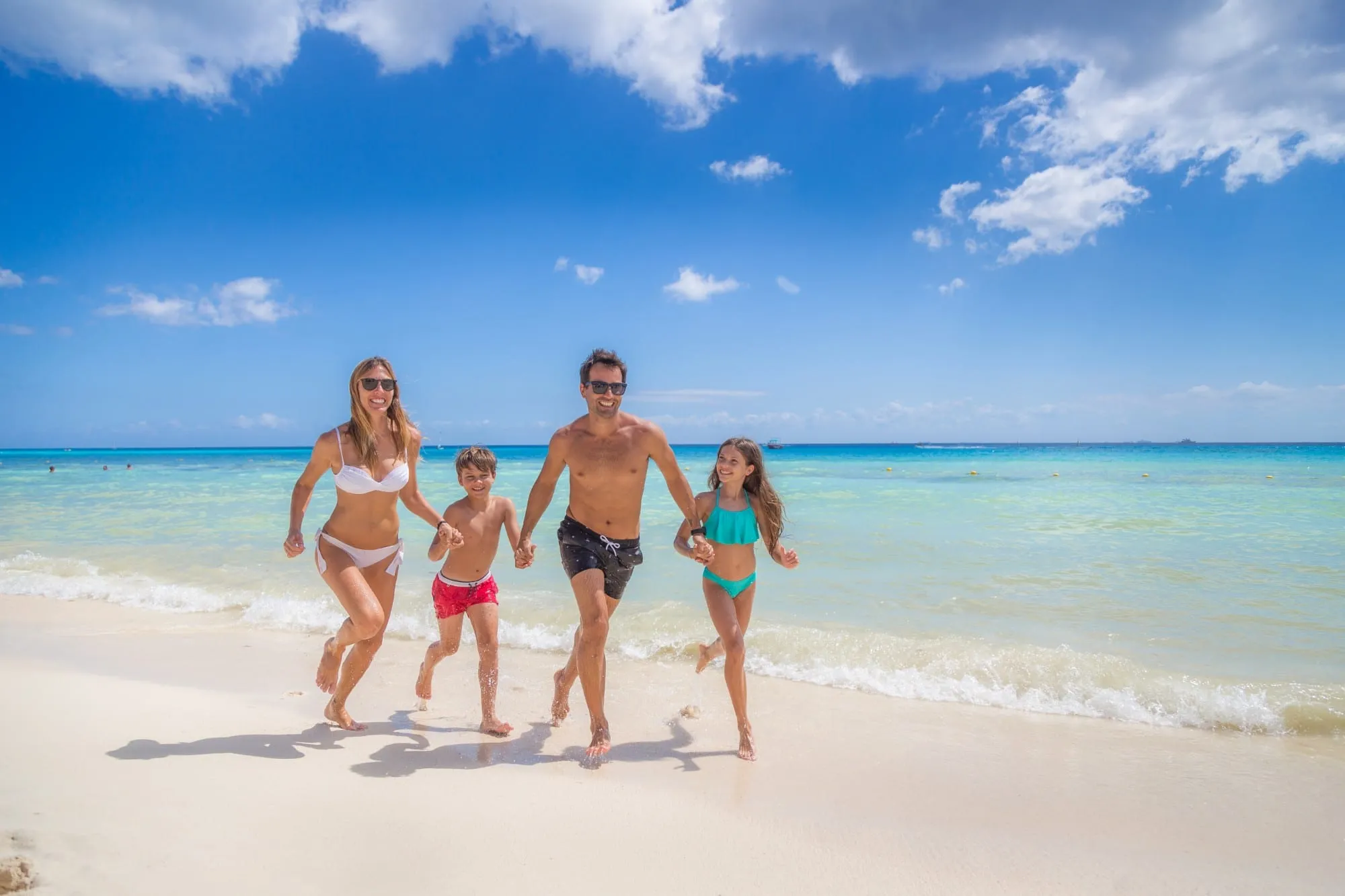 A family of four running hand in hand along a white-sand beach at the water’s edge