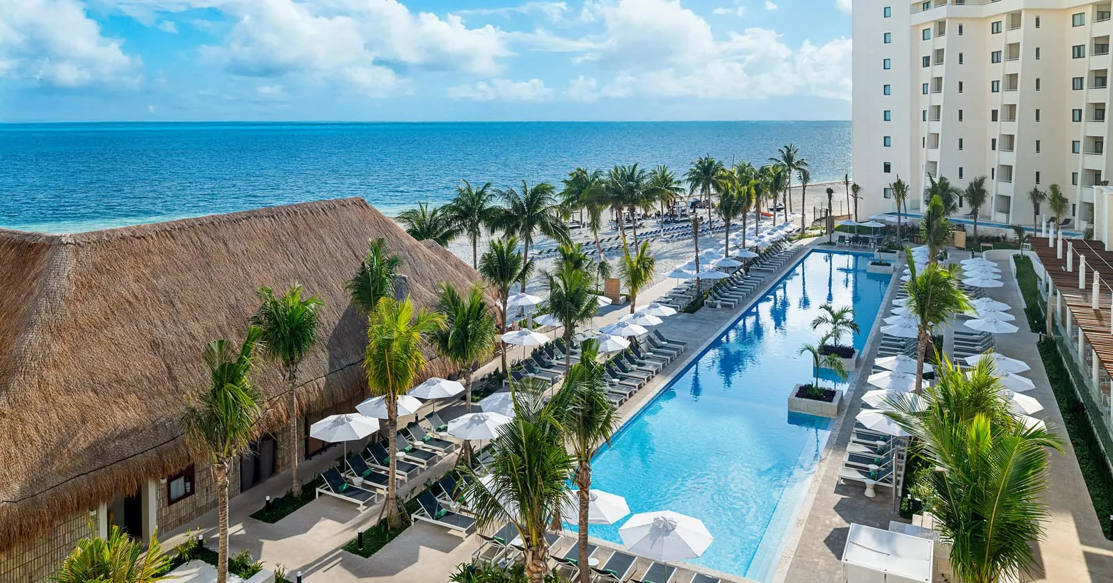An elevated view across a beachfront resort — a thatched palapa roof, a long pool lined with white umbrellas, a guest-room block to one side and open sea past the sand.